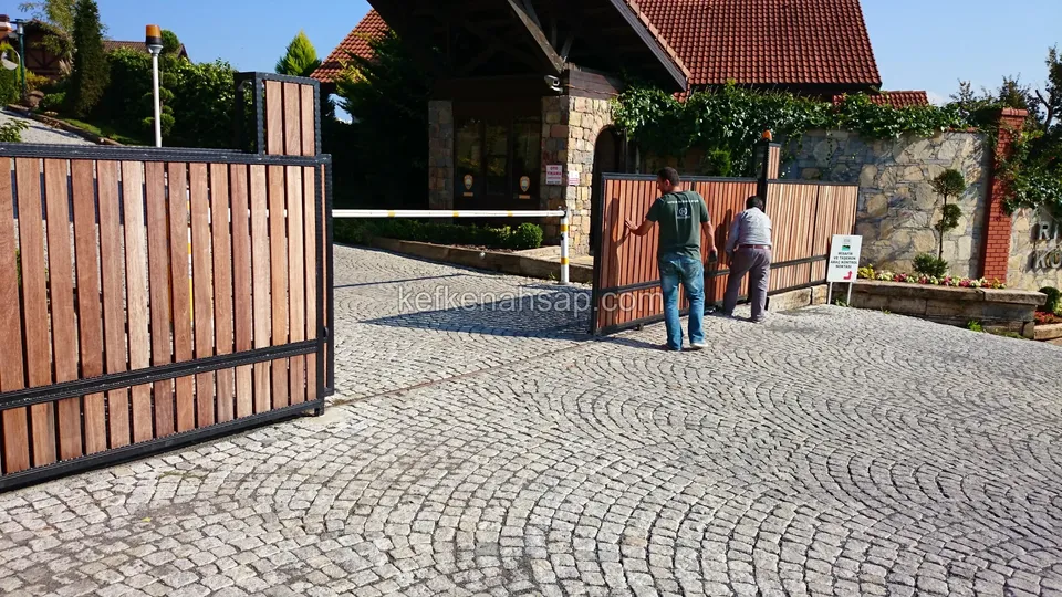 Küp taş döşeli girişte, siyah çerçeveli dikey ahşap çıtalı sürgülü kapının bir kanadı; uzakta kapı önünde iki kişi