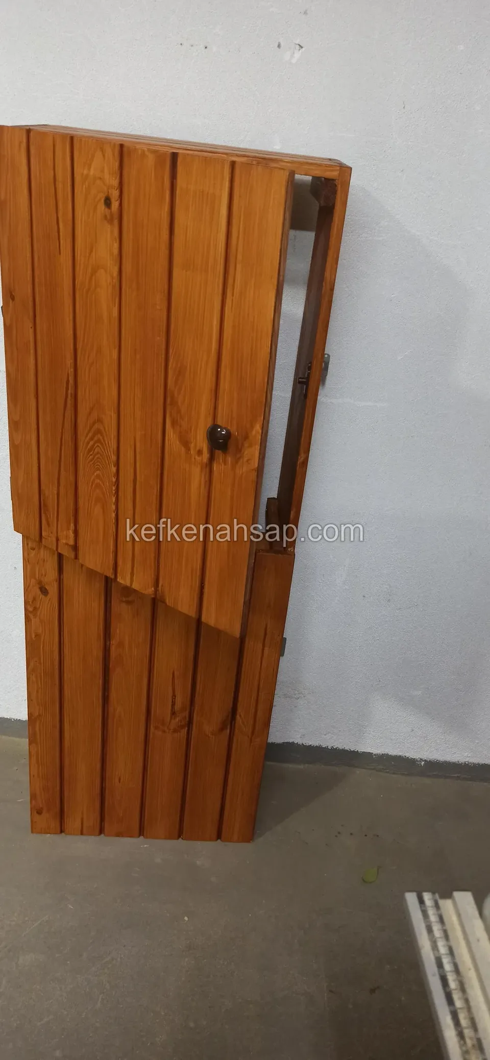 Beyaz duvara yaslanmış, dikey tahtalı ve ortasında küçük bir tutamağı olan açık kahve ahşap kapak iki parça hâlinde