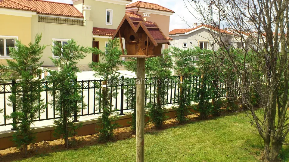 Çimlik bahçede uzun bir direğin tepesine monte edilmiş, bordo çatılı çok bölmeli ahşap kuş evi