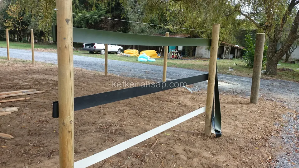 Kalın ahşap direkler arasına gerilmiş siyah ve beyaz bant, arkada çakıl yol ve ağaçların altındaki araçlar