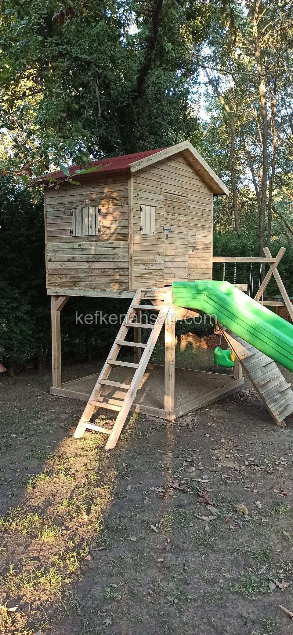 Direkler üzerinde yükseltilmiş kırmızı çatılı ahşap oyun evi; el merdiveni, yeşil kaydırak ve salıncak