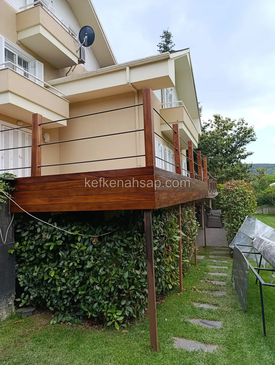 Raised dark red-brown deck platform beside a building, resting on timber posts, with a railing of thin cables