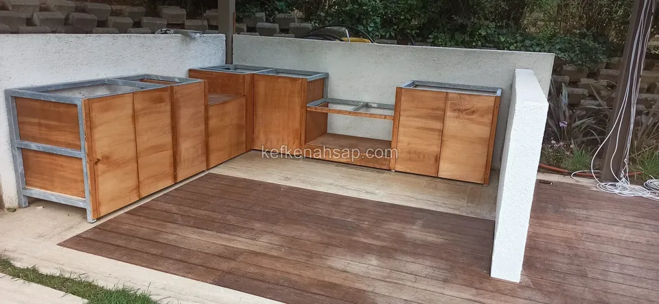 Ahşap deck üzerinde, metal çerçeveli ve ahşap kapaklı L biçimli dış mutfak dolapları
