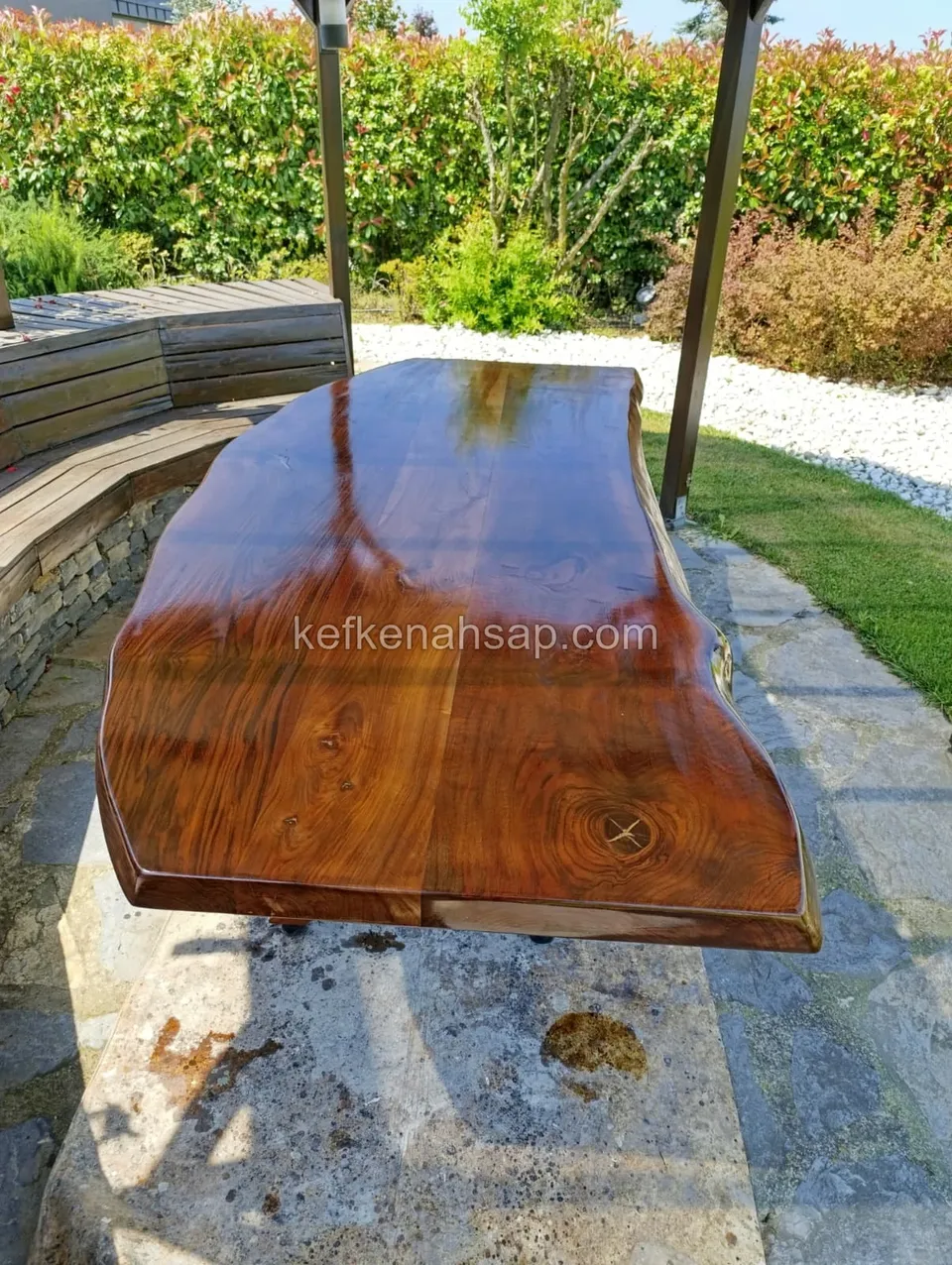 Glossy varnished live-edge solid wood tabletop on a stone patio in a garden, next to a curved wooden bench