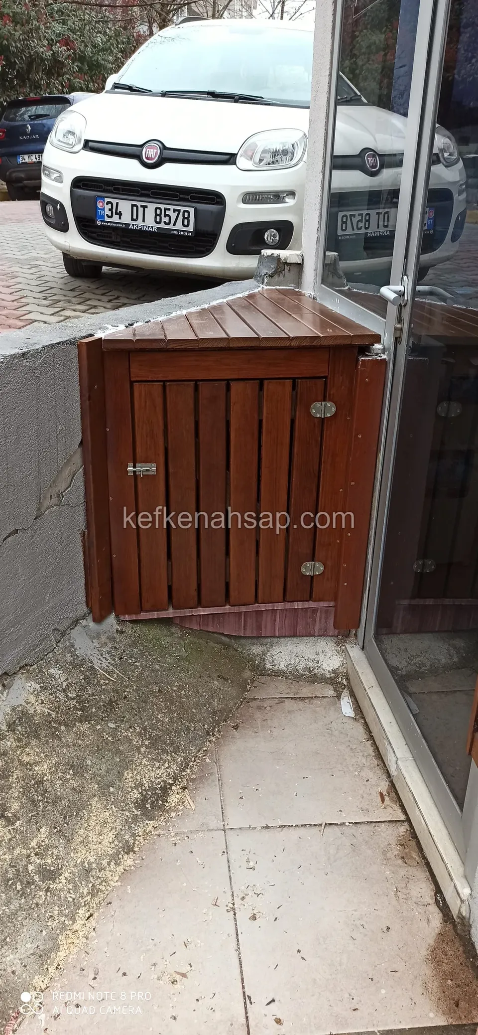 Cam dükkân kapısının yanında, kaldırım köşesine oturtulmuş, üstü eğimli ve önü dikey tahtalı kızıl kahve küçük ahşap dolap