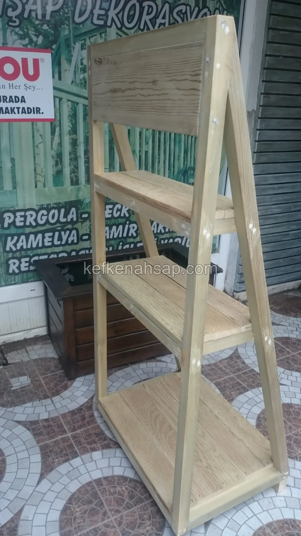 Dükkan önünde duran, A biçiminde, üç katlı açık renk ahşap raf; üstünde koyu tahtalı panel