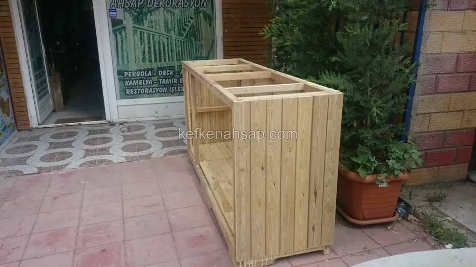 Cam önlü dükkânın girişinde yan yüzeyi dikey tahtalarla kaplanan açık renkli emprenyeli dolap gövdesi, yanında saksı