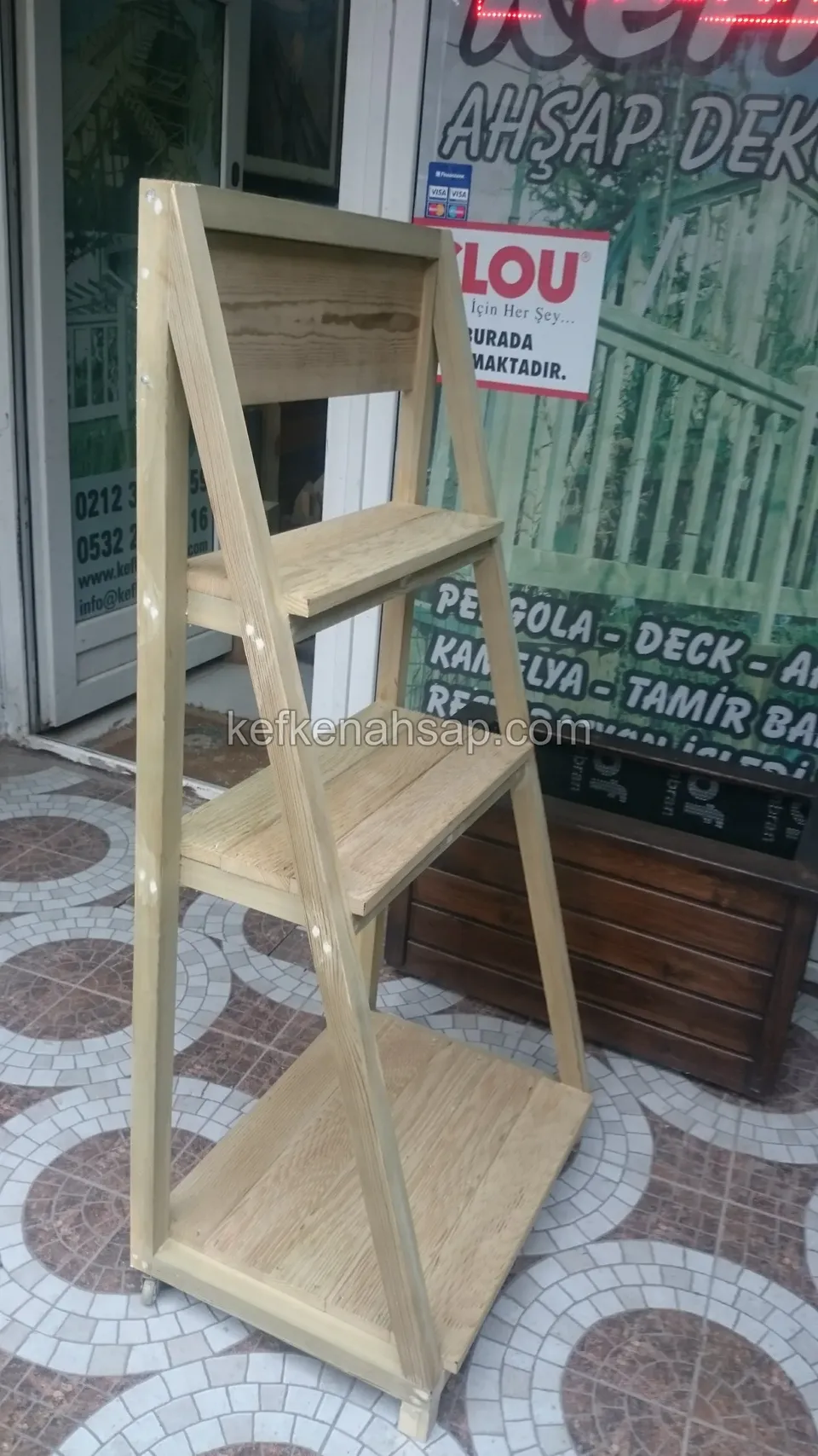 Ein dreistufiges helles Holzregal in Stufenform auf einem Mosaikboden am Ladeneingang