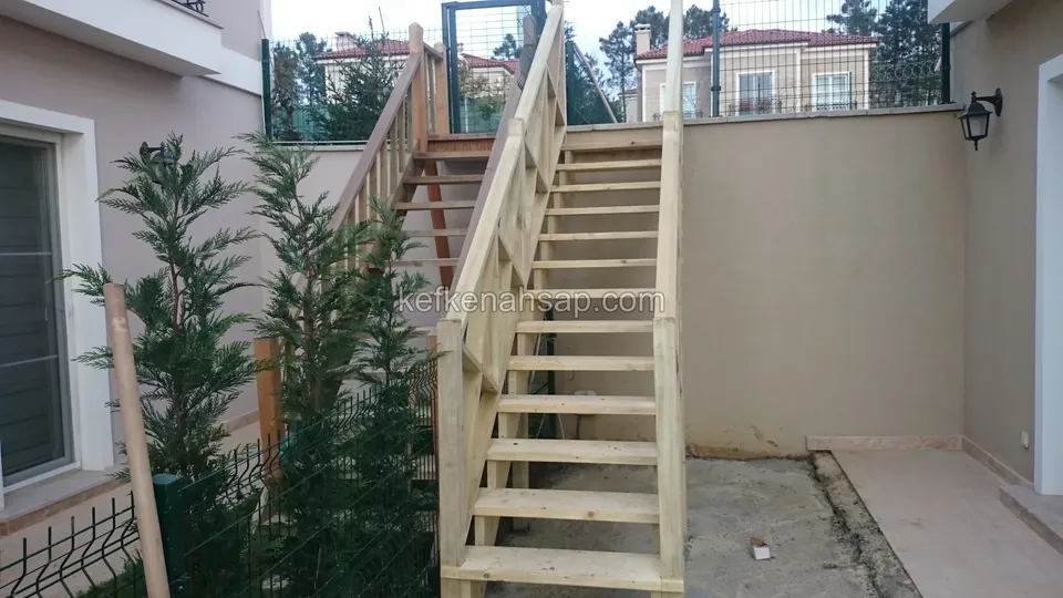 İki duvar arasındaki dar alanda yukarı çıkan ahşap merdiven; solda çalılar ve tel çit, duvarda lamba