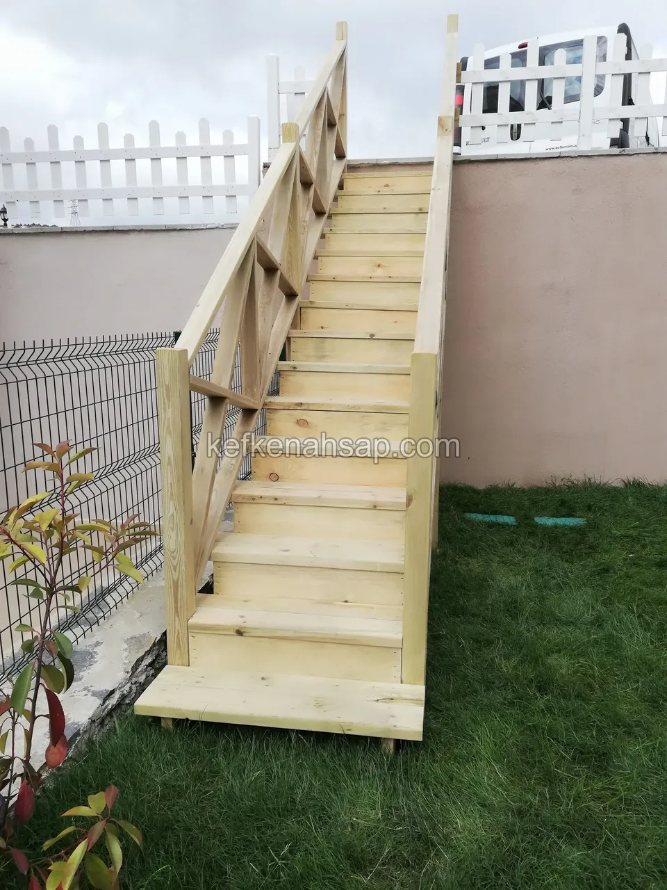 Gerade Holztreppe mit Kreuzstreben-Geländer entlang einer Gartenmauer, oben ein weißer Lattenzaun, links grüner Maschenzaun