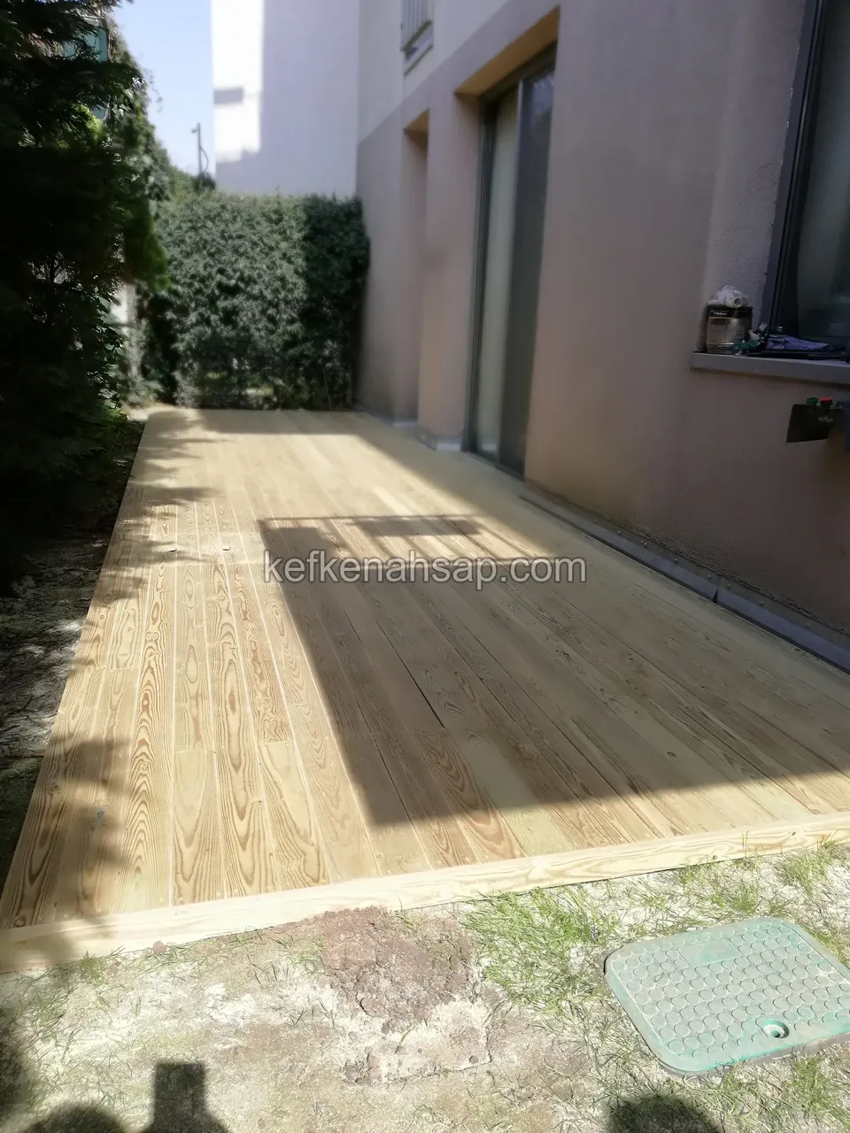Wide light-coloured deck laid in front of a wall, with bare soil around it and a green manhole cover
