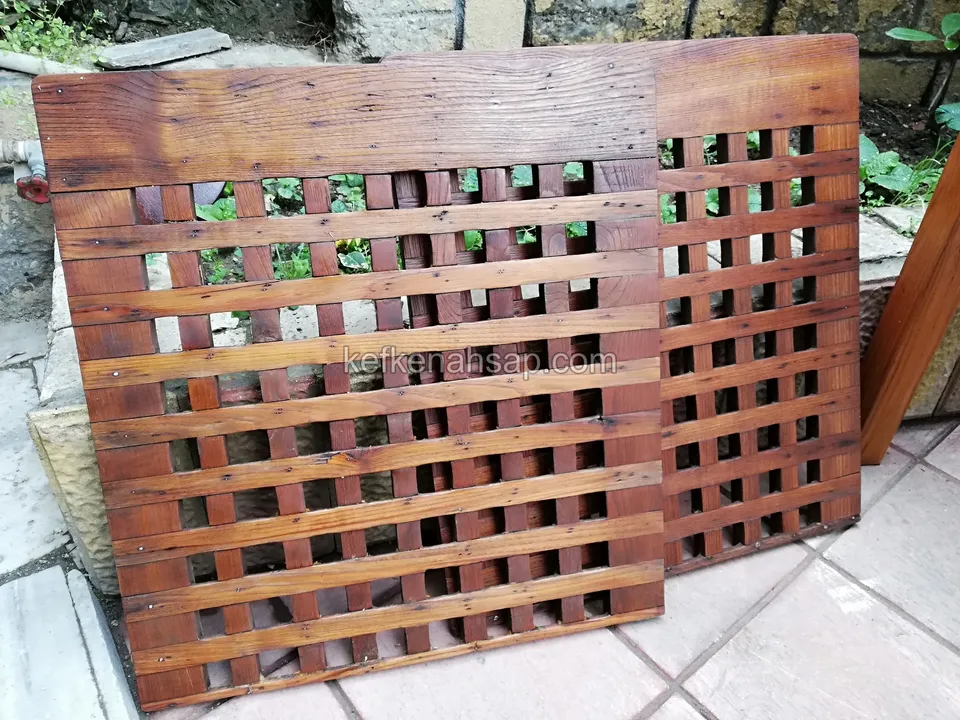 Taş döşemede duvara yaslanmış iki kırmızımsı vernikli ahşap kafes panel; çıtalar çapraz bindirilmiş