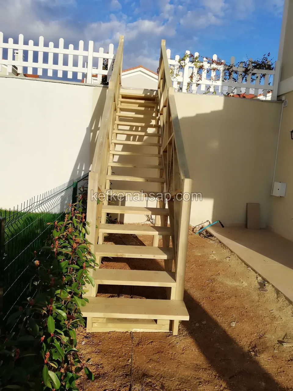 Holztreppe zwischen zwei Mauern, links rotblättrige Sträucher, ganz oben ein weißer Lattenzaun