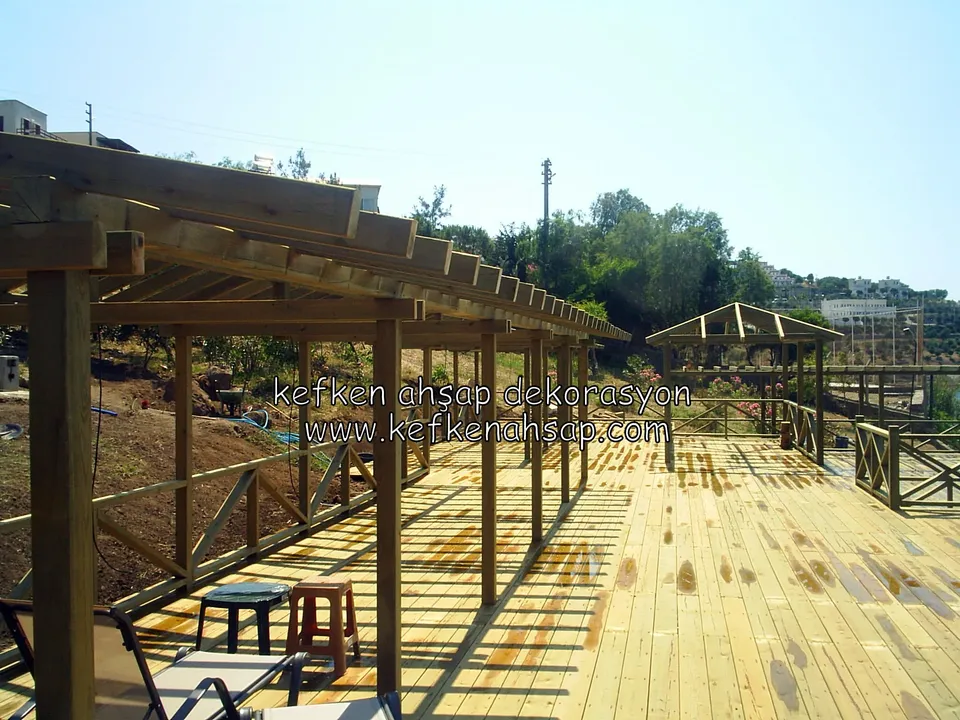Eğimli arazide uzayıp giden ahşap pergola, geniş deck, yan korkuluk, ön tarafta plastik taburelerle çalışma alanı