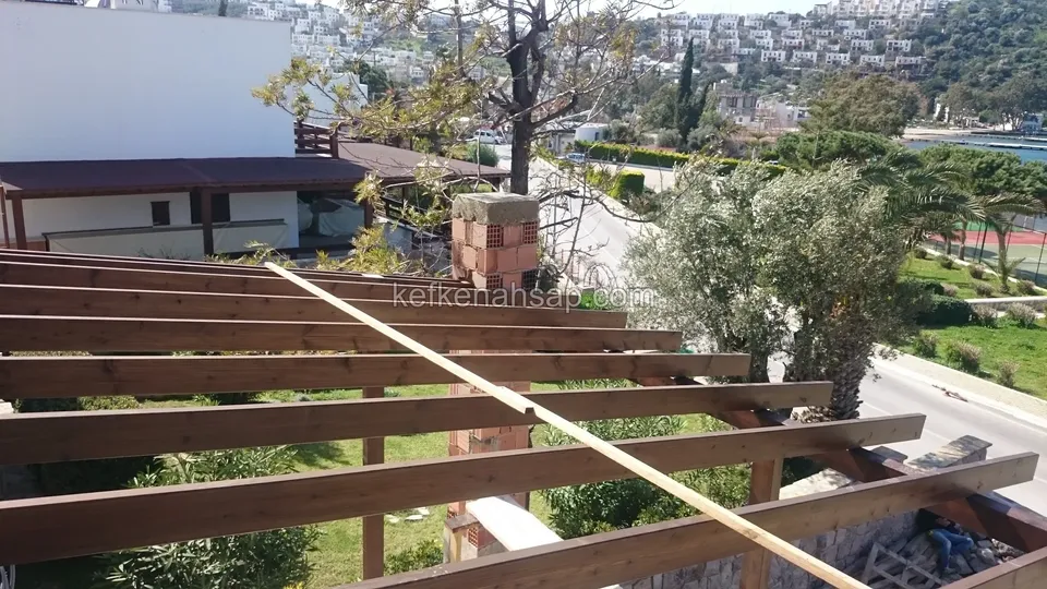 Eğimli arazide yatay vernikli pergola kirişleri, tuğla kolon, zeytin ağacı ve uzakta sahil yerleşimi