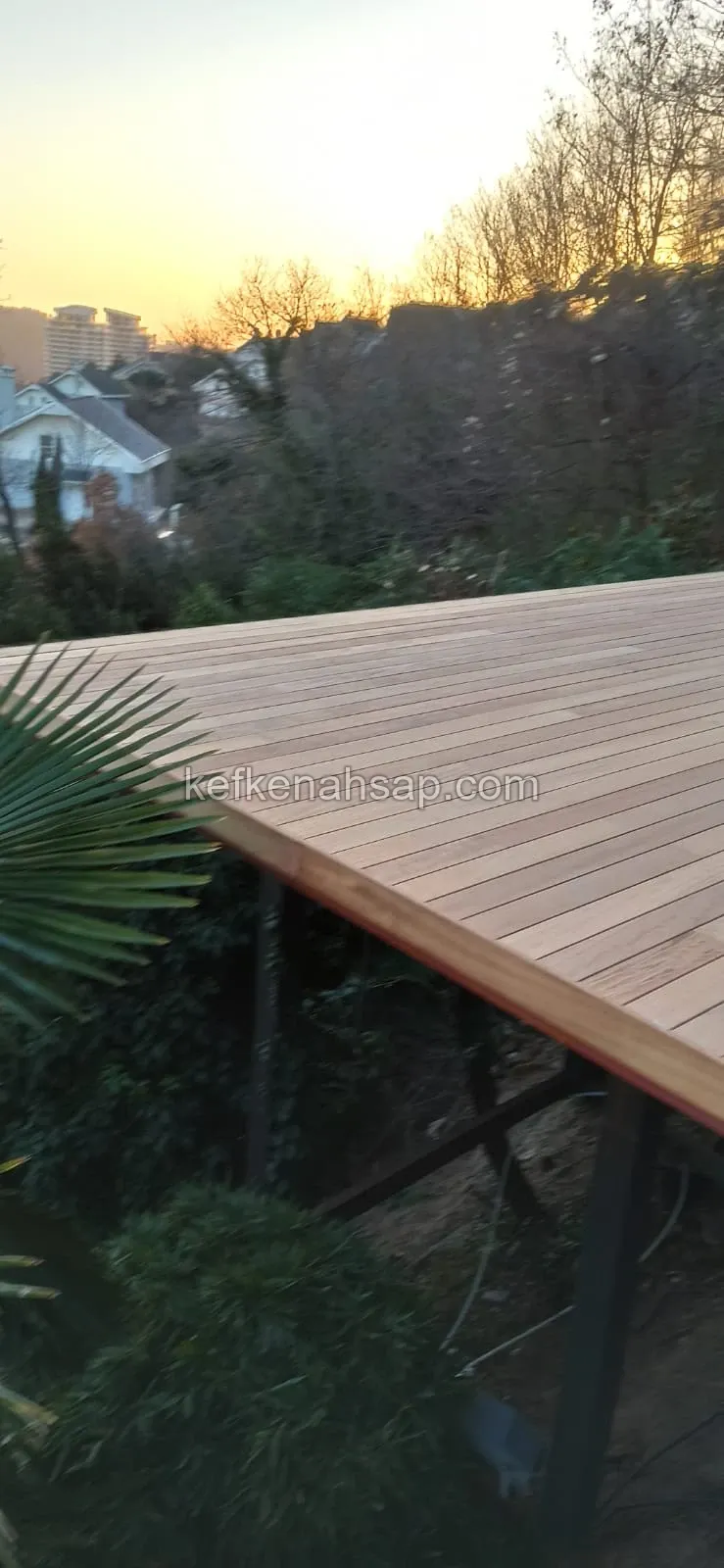 Gün batımında, ağaçlıklı bir yamaca kurulmuş metal ayaklı yükseltilmiş ahşap deck zemini ve arkada evler