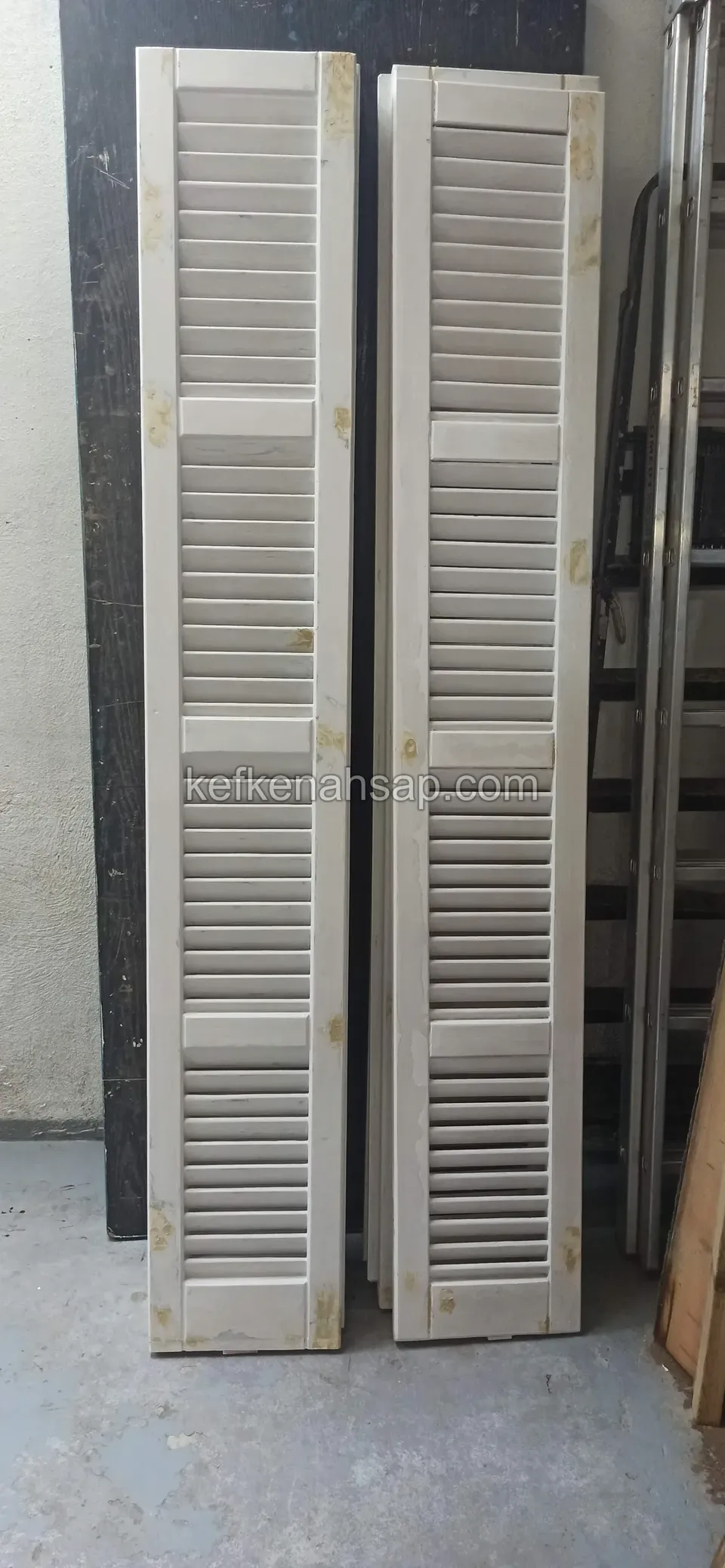 Duvara dayalı, beyaz boyası yer yer dökülmüş iki uzun jaluzi panjur kanadı, yanında merdiven ve koyu bir panel