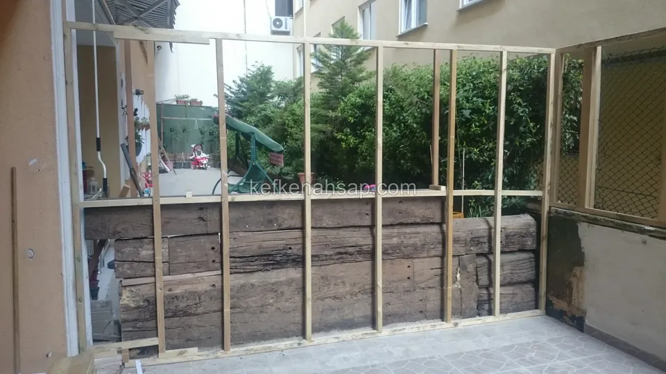 Koyu renkli eski kalın ahşap kirişlerin önünde, ince dikmelerle kurulmuş ahşap duvar karkası; arkada bahçe görünüyor