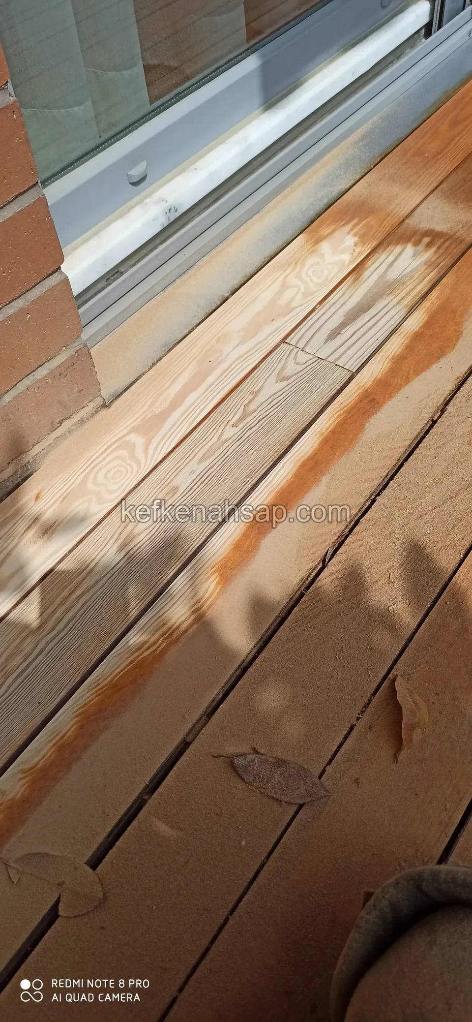 Verwittertes Deck vor einem Glasrahmen mit abblätternder orangefarbener Beschichtung und kahlen Holzstellen