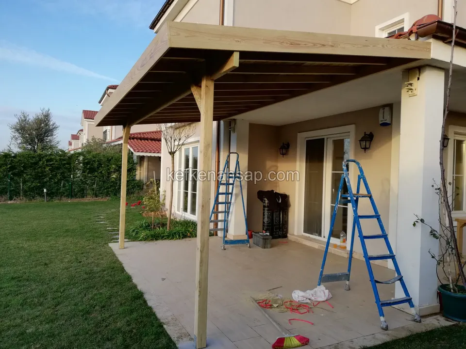 Geneigtes Holzvordach auf schlanken Pfosten am hinteren Eingang eines Hauses, auf der Terrasse zwei Leitern