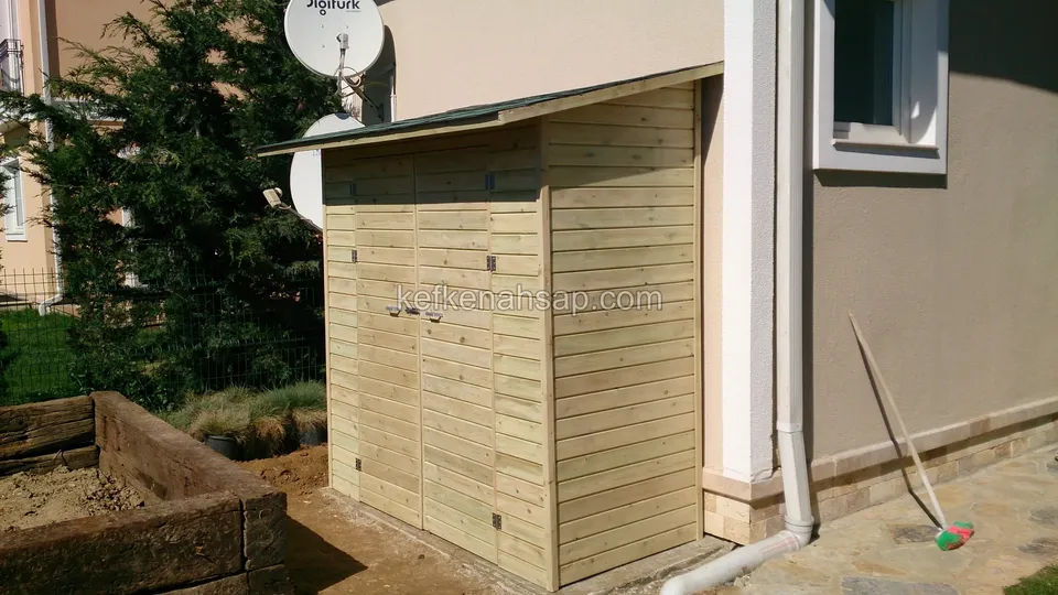 Ev duvarının köşesine yerleştirilmiş yatay tahtalı açık renk ahşap depo ve yanında bahçe yatağı