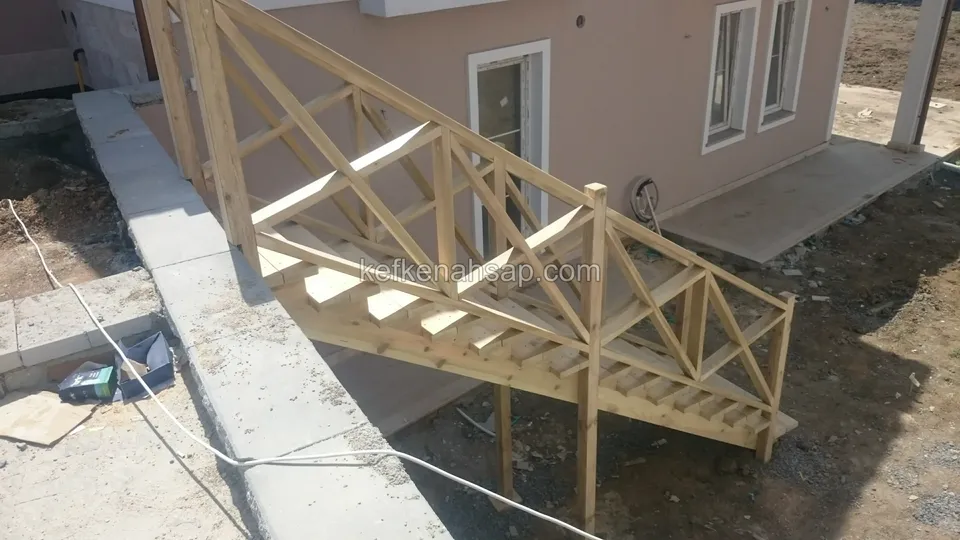 Evin yanında çaprazlı korkuluklu ahşap merdiven; beton kenar, yerde kablo ve açık toprak