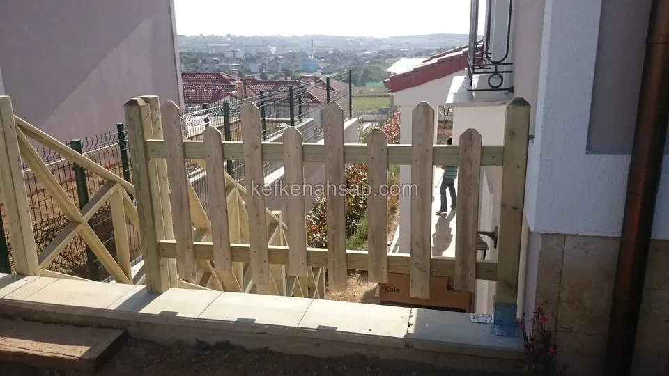 Evin yan duvarında basamak başına konmuş yeşilimsi ahşap piket kapı ve solda X çaprazlı korkuluk, arkada şehir manzarası