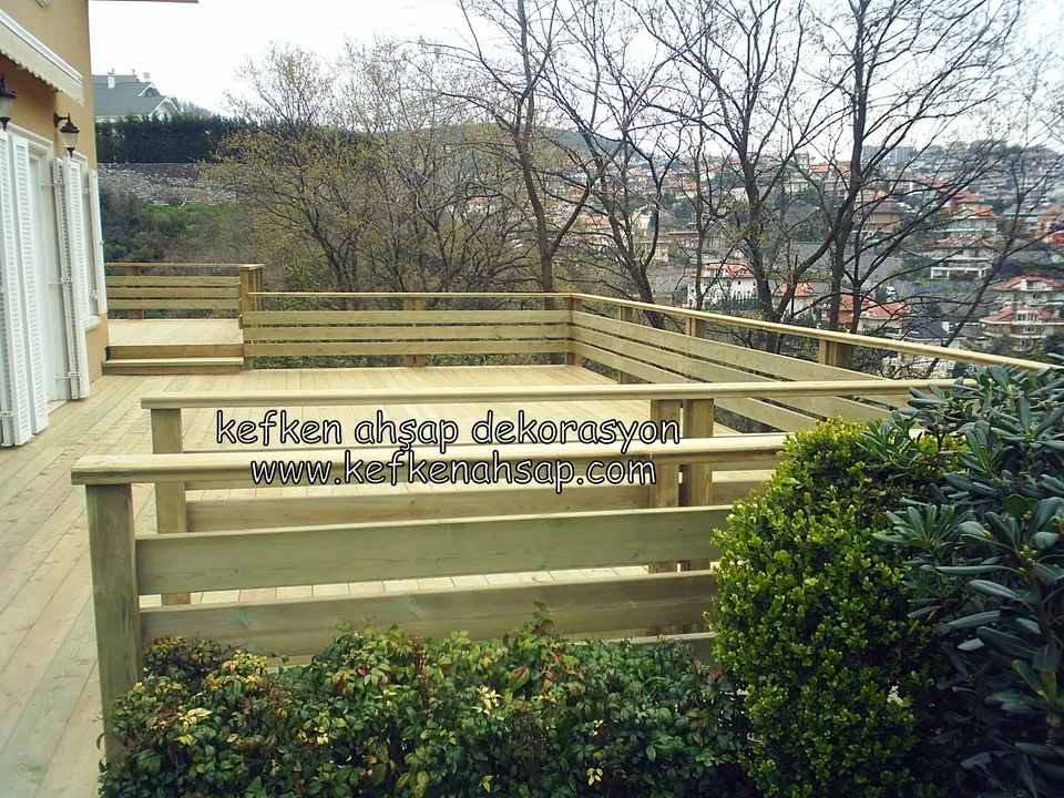 Evin camlı kapısının önünde L biçimli açık tonlu deck, çevresinde yatay tahtalı korkuluk ve yeşil çalılar, arkada ağaçlar