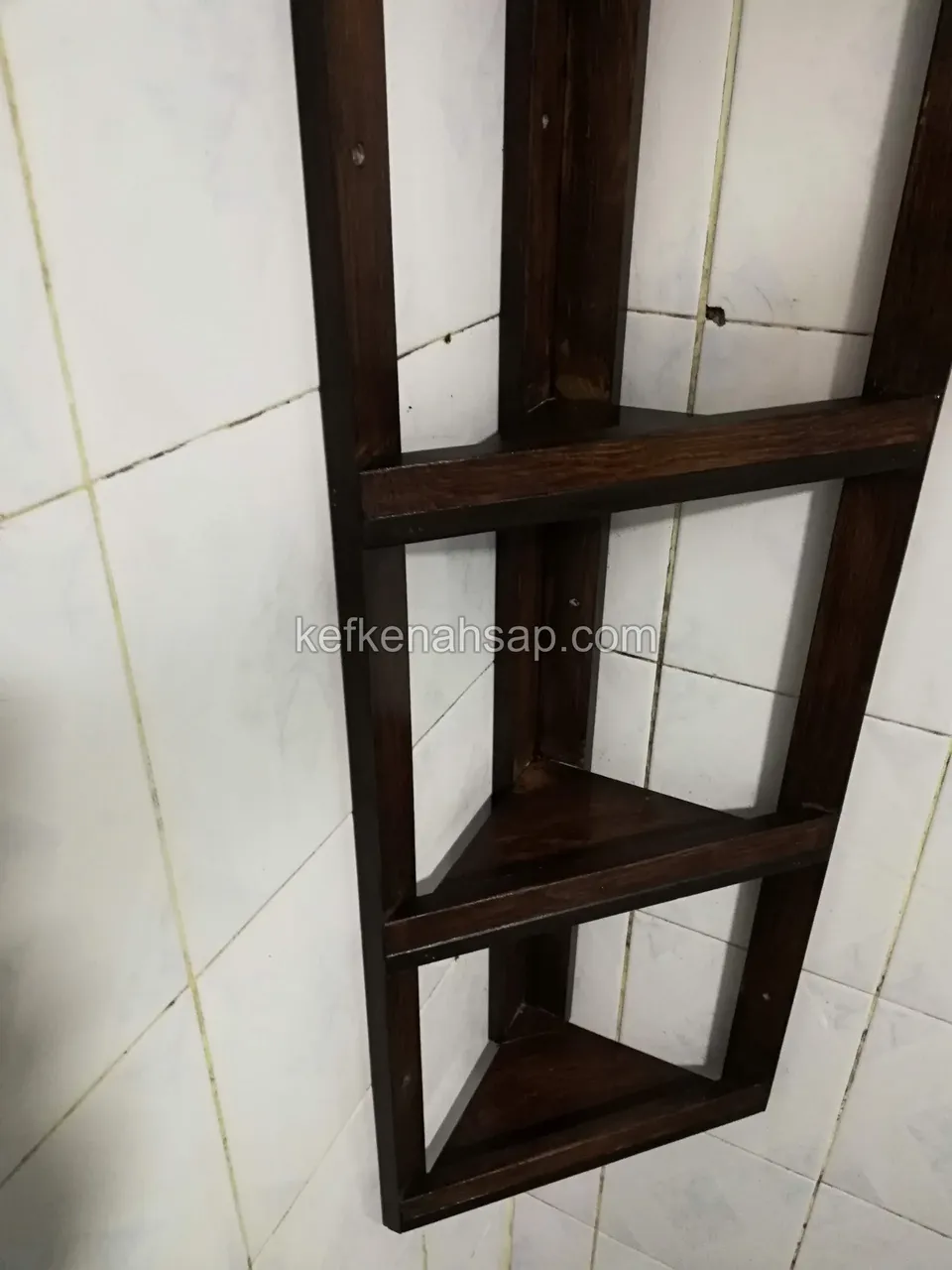 Beyaz fayans duvarda koyu vernikli ahşap köşe rafı; üç katlı, ince çıtalı yan kenarlı