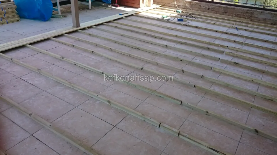 Fayans döşeli zeminin üzerine eşit aralıklarla sıralanmış ince emprenyeli çıtalar, arka planda cam kapılı teras