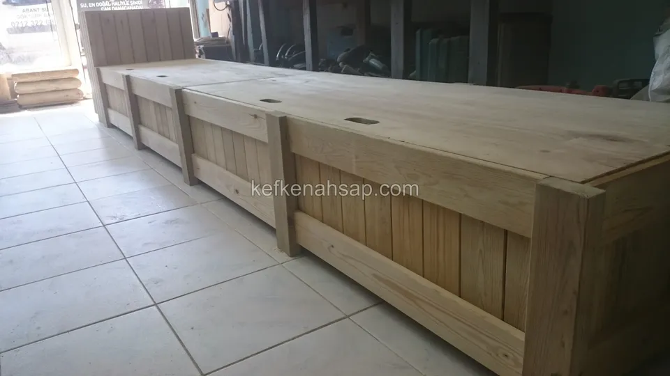 Eine lange Truhenbank aus hellem Holz mit drei Deckeln entlang einer Wand auf einer Terrasse mit hellen Keramikfliesen
