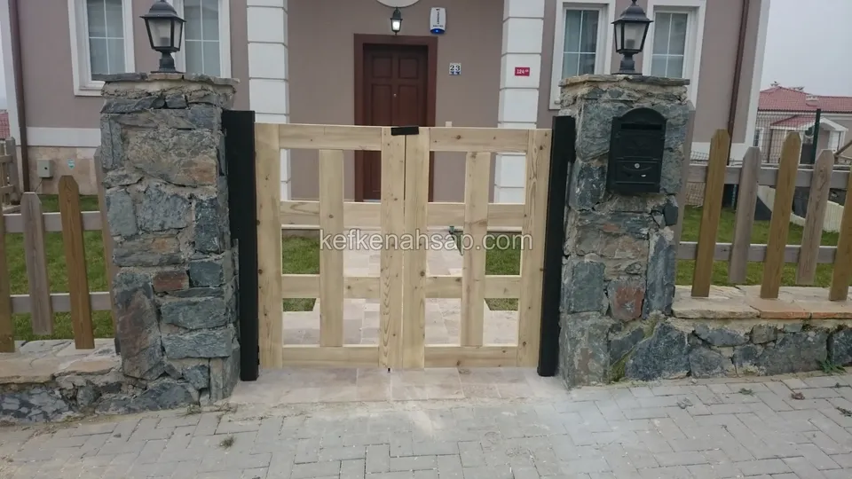 Fenerli taş kolonlar arasında iki kanatlı açık renk ahşap kapı ve iki yanında ahşap çit