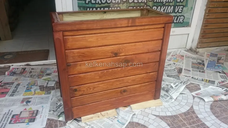 Dükkân önünde gazete serili zeminde duran, yeni vernik sürülmüş turuncu-kahve yatay tahtalı ahşap çiçeklik