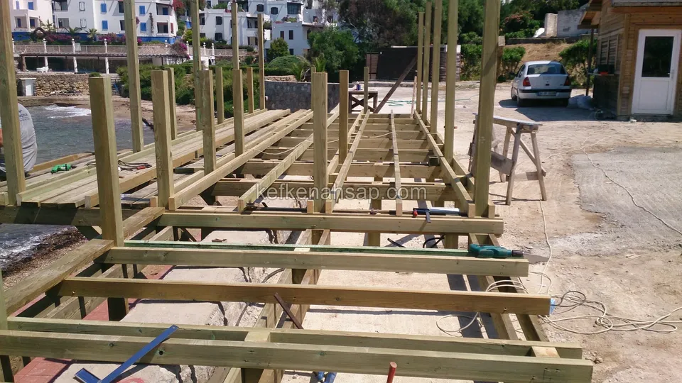 Wide timber sub-frame covering a long area by the sea, with rows of posts, and white buildings and vehicles behind
