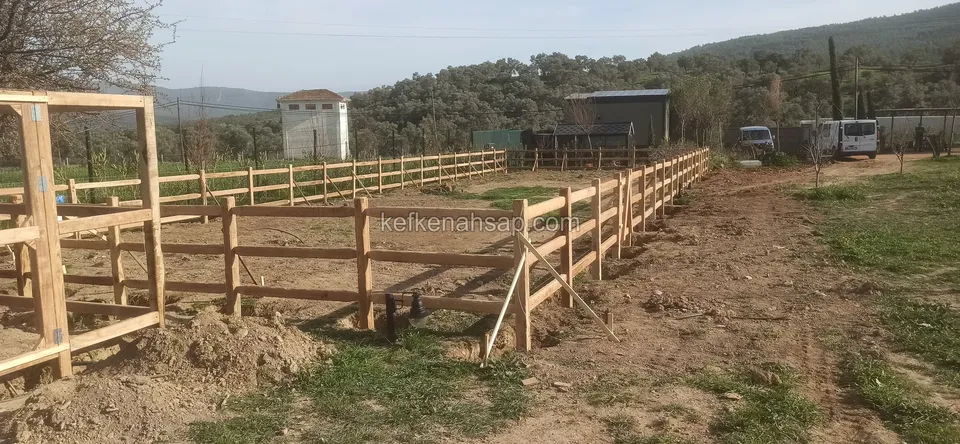Geniş açık arazide uzanan yatay çıtalı çit, önde ahşap kapı çerçevesi, arka planda tepeler ve birkaç bina