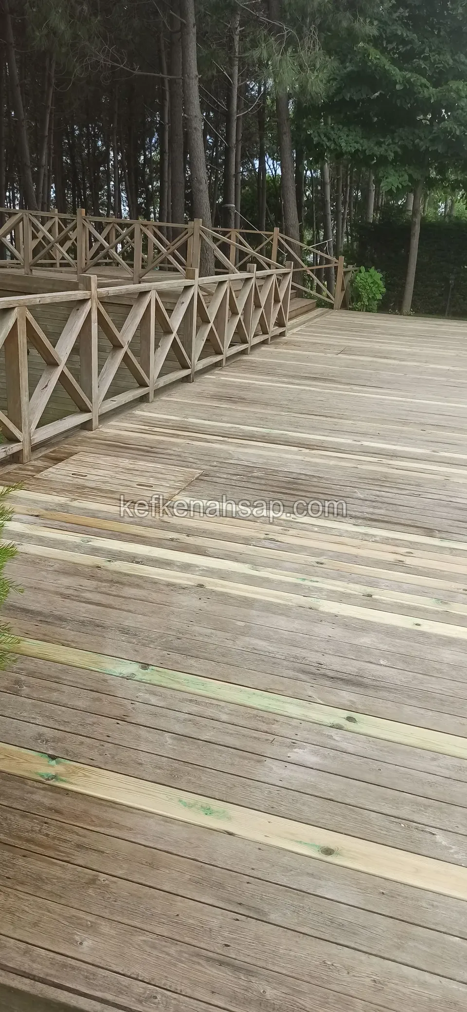 Broad deck with a cross-braced railing and new pale boards among old grey ones, with pine trees behind