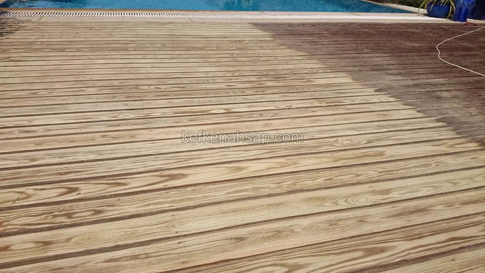 Havuz kenarındaki geniş deck yüzeyinin sağ bölümü koyu bir renkle işlenmiş, sol bölümü ham tahta; uzakta havuz ve ip