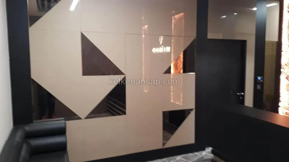 Siyah çerçeveli, bej parlak yüzeyli ve üçgen boşluklu geometrik duvar paneli; yanında siyah deri koltuk var
