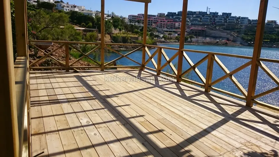 Göle bakan, çapraz çıtalı ahşap korkuluklu açık renkli deck; tahta yüzeyde korkuluk ve direk gölgeleri