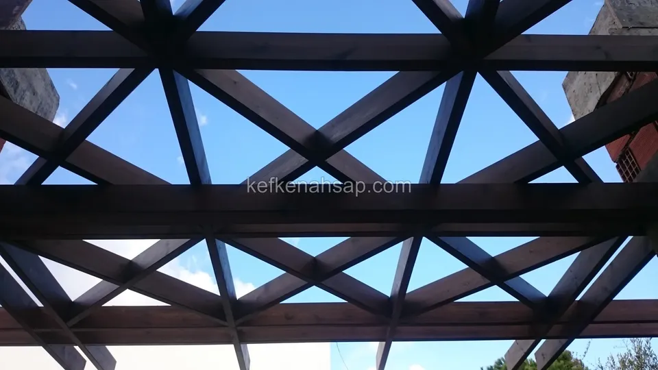 Alttan görünen koyu vernikli üçgen desenli pergola kafesi, aralardan mavi gökyüzü, köşede tuğla kolon