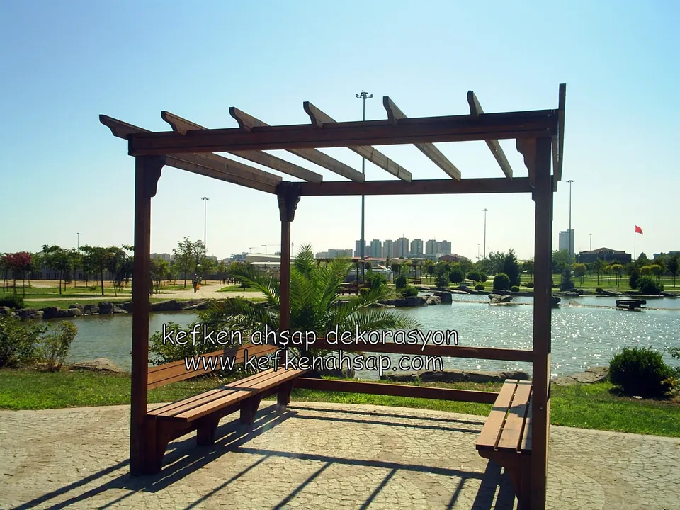 Göl kenarındaki parkta koyu vernikli ahşap pergola, köşede L biçimli bankları ve taş döşeme; arkada göl ve uzak binalar