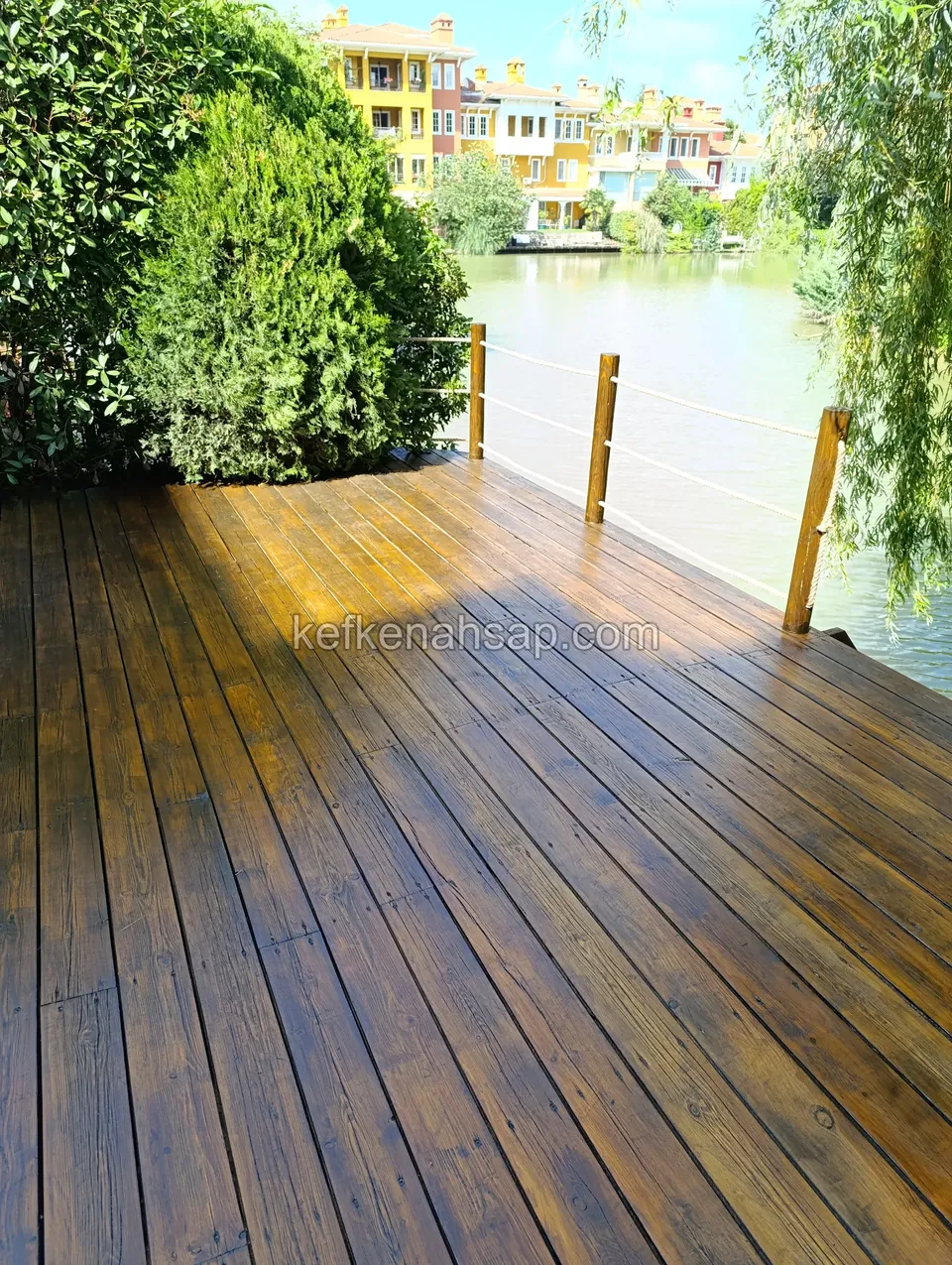 Wet, shiny polished dark brown deck facing a lake, with a rope railing at its edge and colourful houses on the far shore