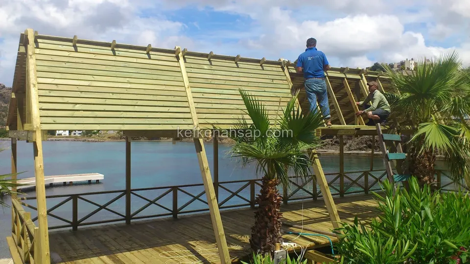 Su kenarındaki deck üzerinde emprenyeli çam pergola çatısı çıtalarını çakan iki kişi, çevrede palmiyeler