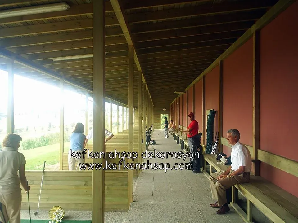 Golf atış alanında ahşap kolonlu, ahşap tavanlı uzun sundurma; kırmızı duvar önünde bank, oyuncular ve golf topları