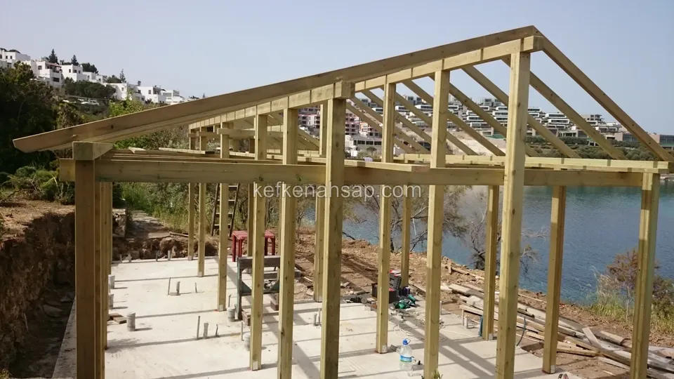 Beton zeminin üzerinde dik direkler ve çatı kirişlerinden oluşan ahşap iskelet, arkada su ve beyaz evler
