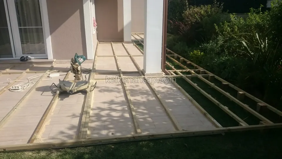 Teras girişinde çimenin kenarında kurulu deck çerçevesi, ortada gönye testeresi ve sağda ayaklara oturan ek kollar