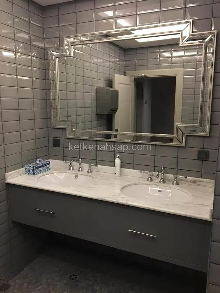 Gri metro fayanslı banyoda, mermer tezgahlı ve iki lavabolu gri boyalı askılı dolap; üstünde çerçeveli geniş ayna