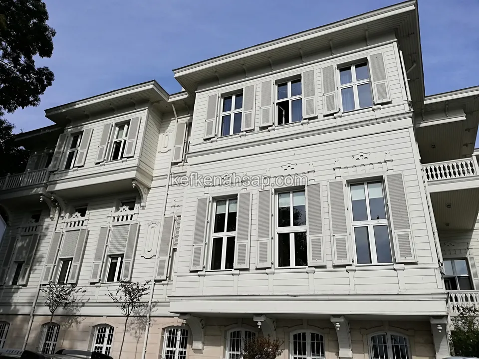 Gri boyalı yatay ahşap kaplamalı, pencereleri kepenkli, üst kenarı işlemeli çok katlı eski tip bir bina cephesi