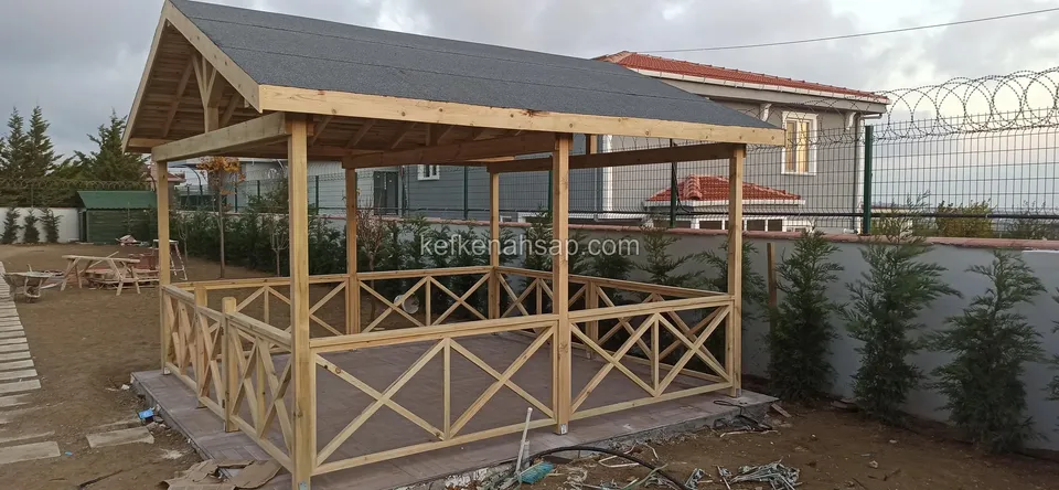 Gri çatı kaplı ahşap kamelya, çapraz çıtalı korkuluk, arkada ev ve servi sırası boyunca çevre duvarı