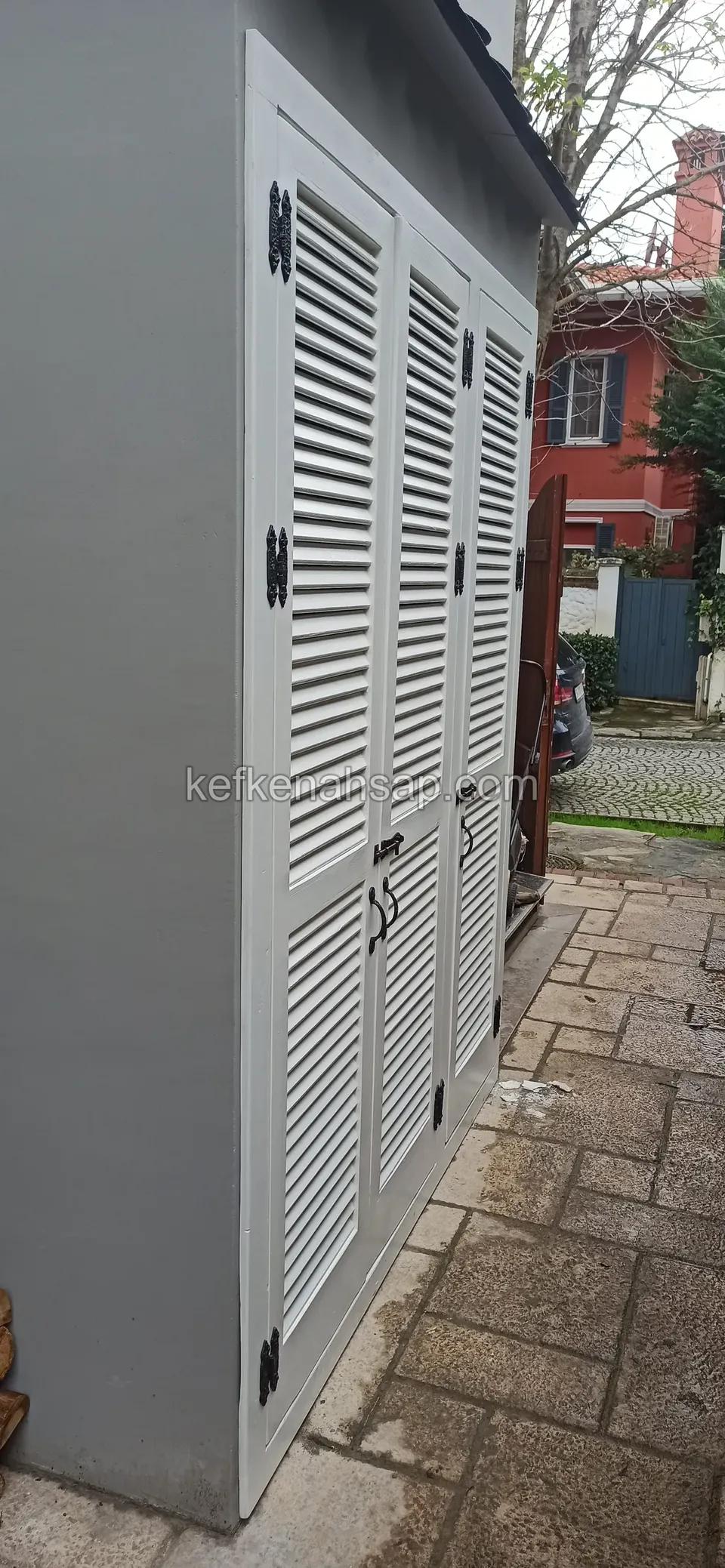 Ein hell gestrichener Holzschrank für draußen mit Lamellentüren neben einer grauen Wand, auf Steinpflaster